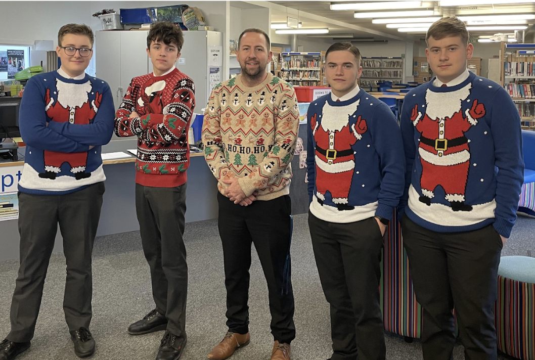 Students and a teacher stood wearing Christmas Jumpers in a library